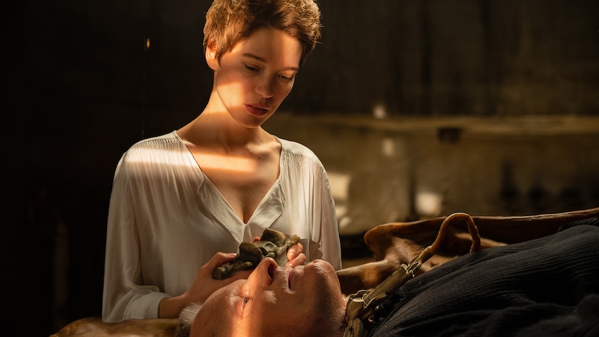 Blonde white woman wears white blouse in darkened room and holds odd object beside a man lying on an operating table.