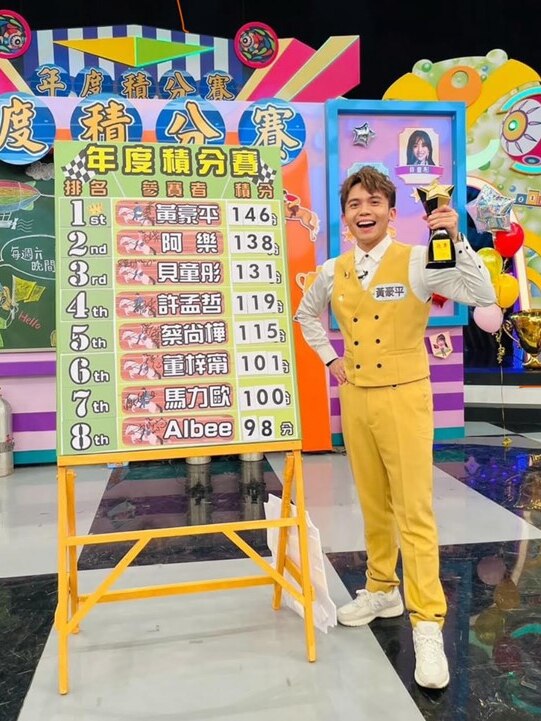 A man dressed smartly in a yellow waistcoat stands smiling next to a large sign on a TV set.