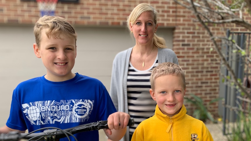 A mother and her two sons stand with a bicycle in the driveway of their home where they have been playing. 