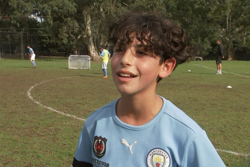 A young boy wearing a blue sports tshirt with logos on it stands with a soccer pitch with players on it behind him