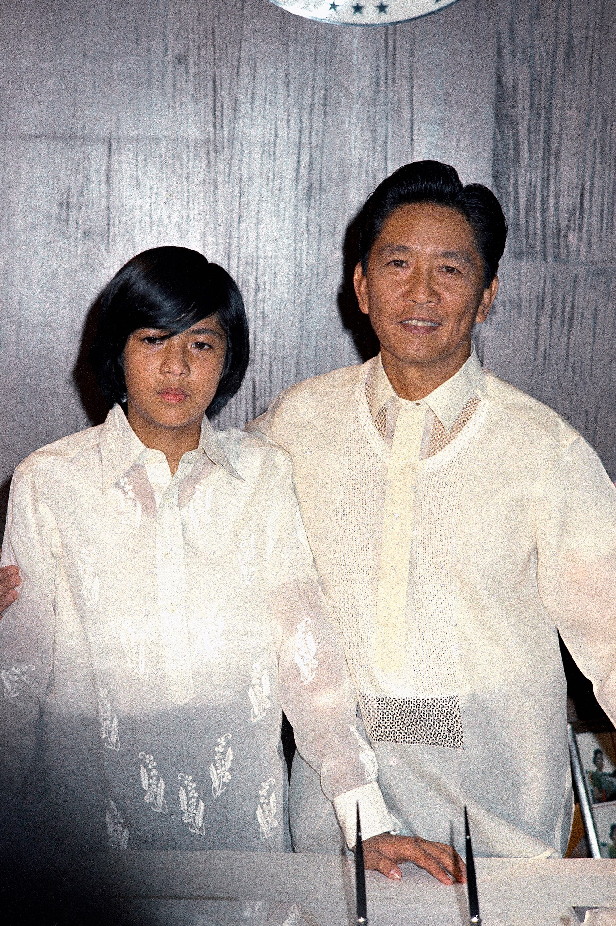 A young Bongbong Marcos in a white shirt standing next to his father, also dressed in a white shirt 