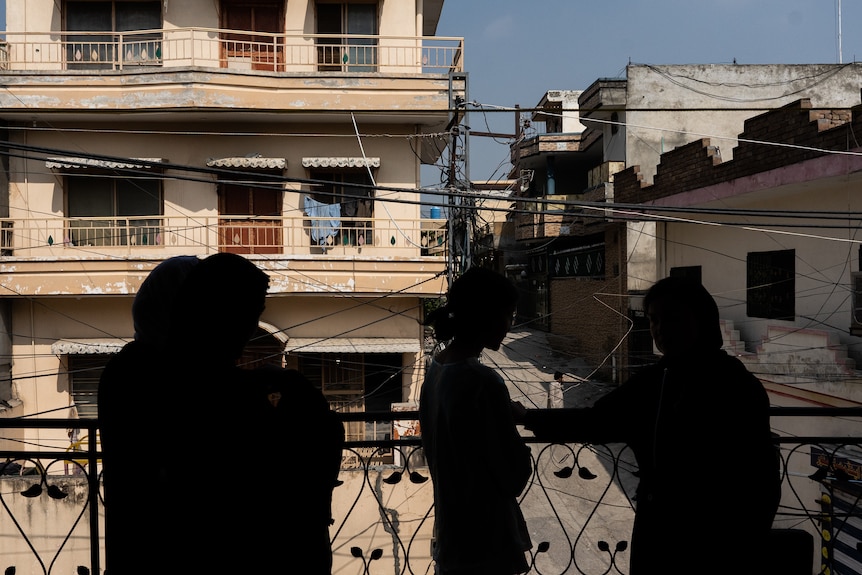 Three people are silhouetted against a backdrop of the street as they stand on the balcony. 