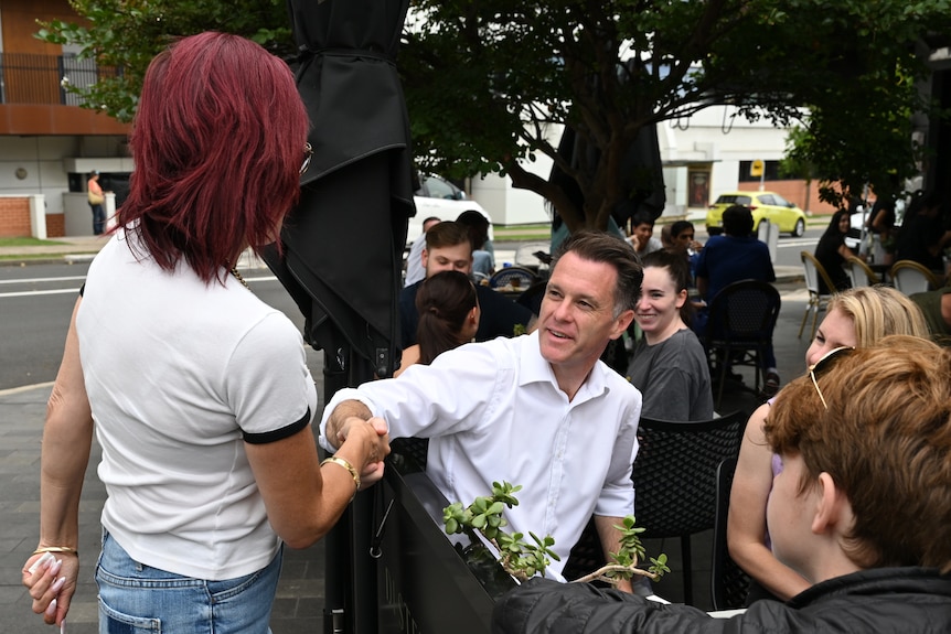 a man sitting at a cafe shaking the hand of another person as they walk past