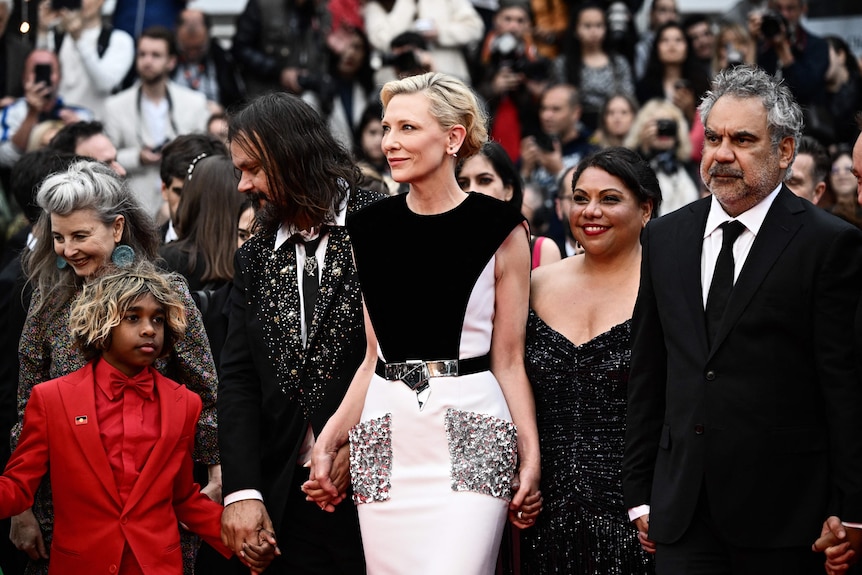 A group of Indigenous and non-Indigenous people on a red carpet, wearing fancy black tie clothing.