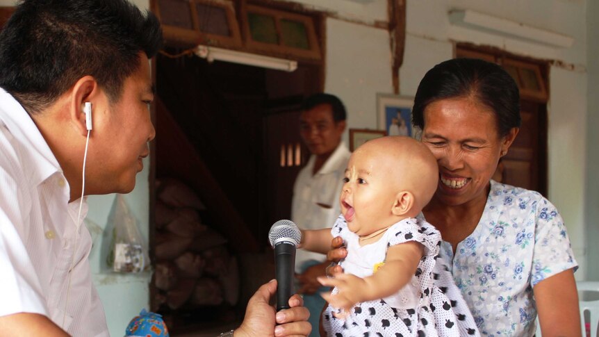 Man with microphone 'interviewing' baby with mum holding her up and smiling.