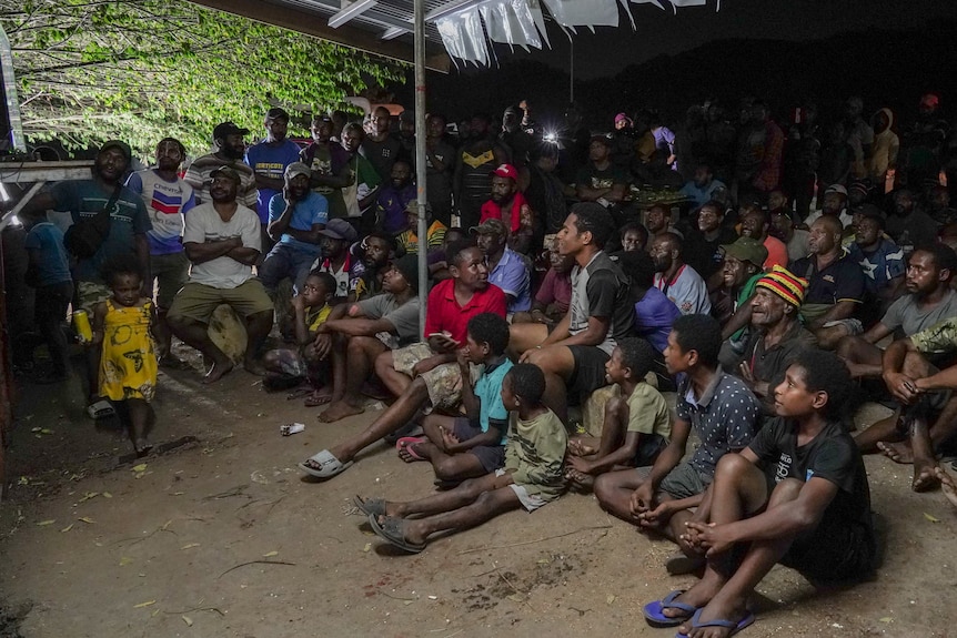 A crowd of people watch a television at night