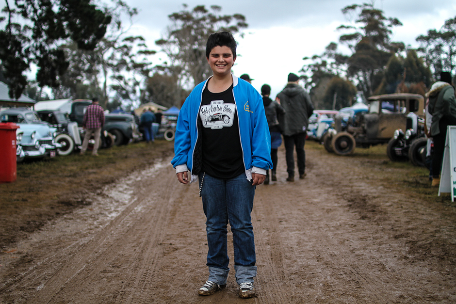A boy with an old-fashioned duck's-tail hairdo stands in the middle of a muddy track, with vintage cars in the background.