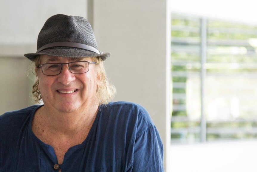 A middle-aged man with curly blonde hair, glasses and a black hat smiles at the camera. 