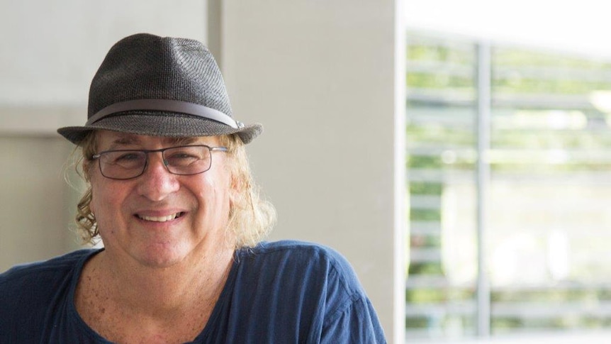 A middle-aged man with curly blonde hair, glasses and a black hat smiles at the camera. 