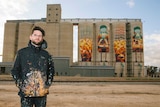 Kyle Hughes-Ogders stands in a paint-splattered jacket in front on the silos he painted.