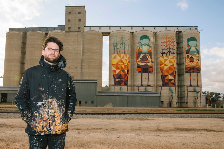 Kyle Hughes-Ogders stands in a paint-splattered jacket in front on the silos he painted.