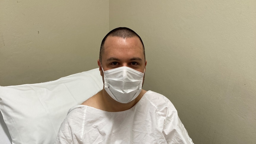 An image of Robbie Barnes in white hospital gown, white face mask, thumbs up in hospital bed