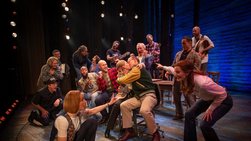 The Australian cast of Come From Away on stage at varying levels; one couple is kissing in the middle of the group.