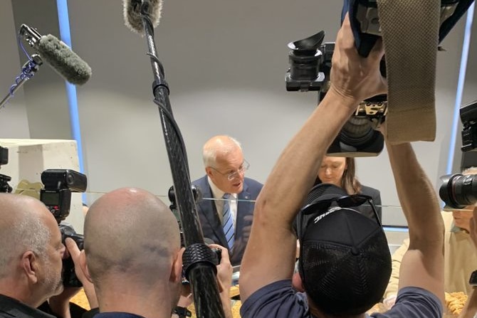Cameraman holding camera above his head trying to get an aerial shot of prime minister talking to a woman in a bakery.