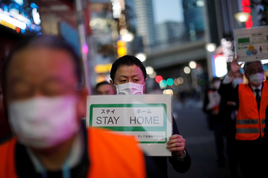 Lines of people walk in the street wearing face masks and holding signs telling people to stay home.