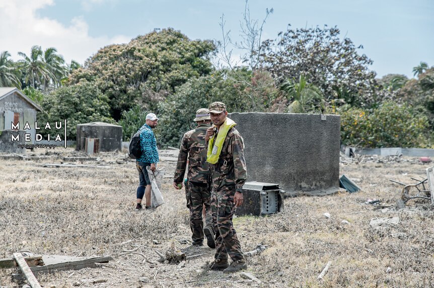 Man, in army uniform, stands infront of tree and house structure on island in Tonga. 