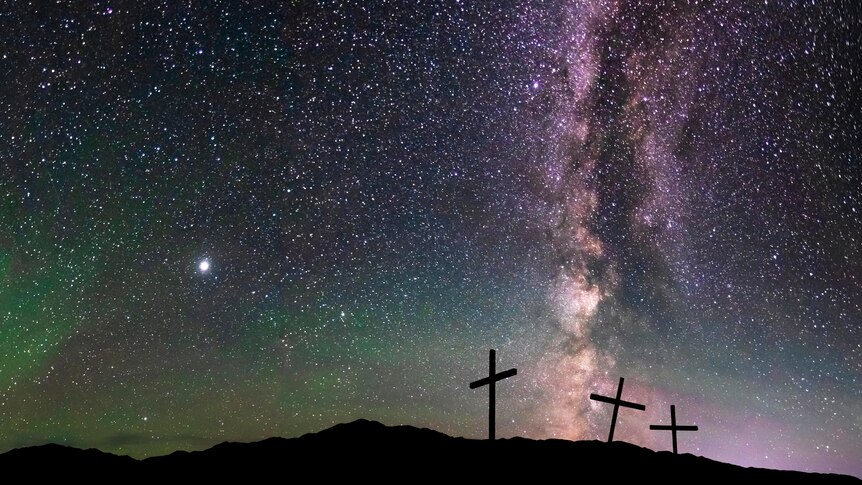 The silhouettes of three crucifixes on a hill stand against a star-filled night sky.
