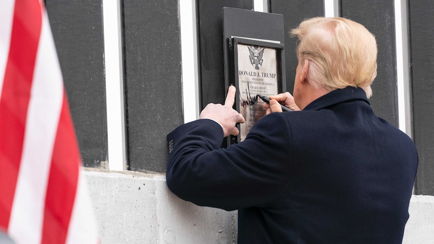 Donald Trump signs a plaque on a fence, his back to the camera.