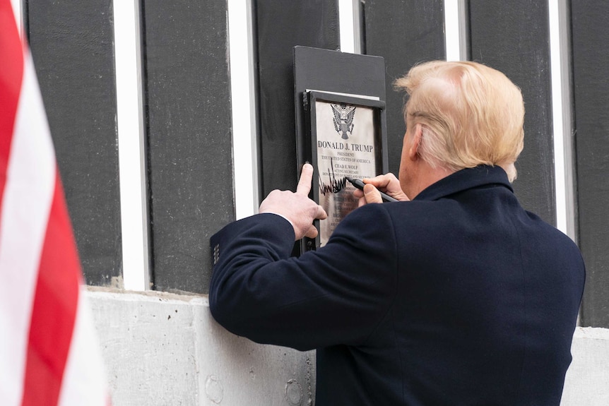 Donald Trump signs a plaque on a fence, his back to the camera.