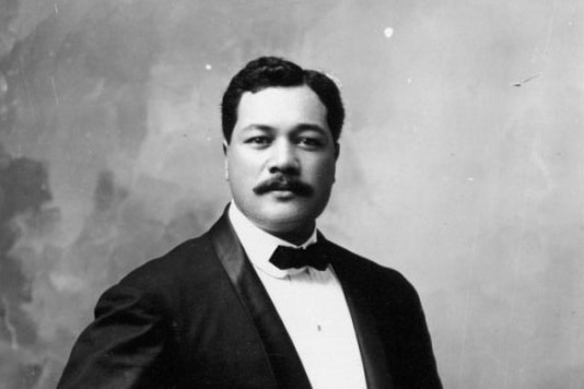 Black and white photo of Ernest Kaʻai, standing wearing suit and bow tie, and holding ukulele, smiling slightly.
