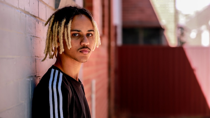 Young man with dark skin and blonder-dyed dreadlocks stands in alleyway with back against orange wall