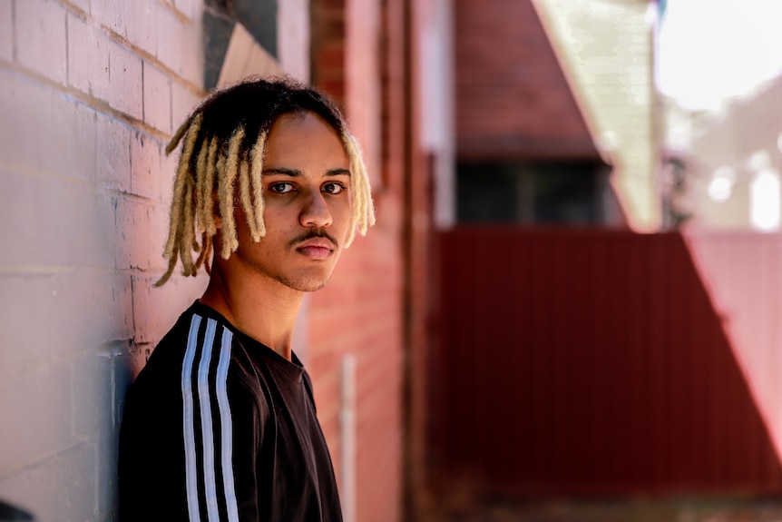 Young man with dark skin and blonder-dyed dreadlocks stands in alleyway with back against orange wall