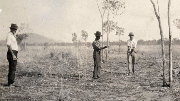 A black and white photo of three men in hats, two holding rods, in a dry-looing field with sparse, scraggly trees.