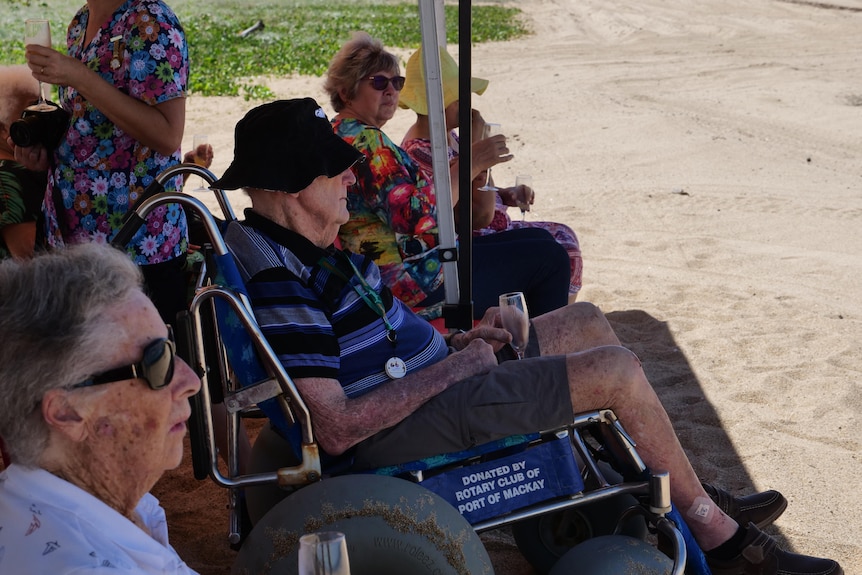Elderly people sit under a shade tent on the beach drinking juice out of champagne glasses. 