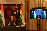 Tehan, suited, sits at a desk in front of Australian and India flag. Morrison appears on a TV to his left.