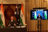 Tehan, suited, sits at a desk in front of Australian and India flag. Morrison appears on a TV to his left.