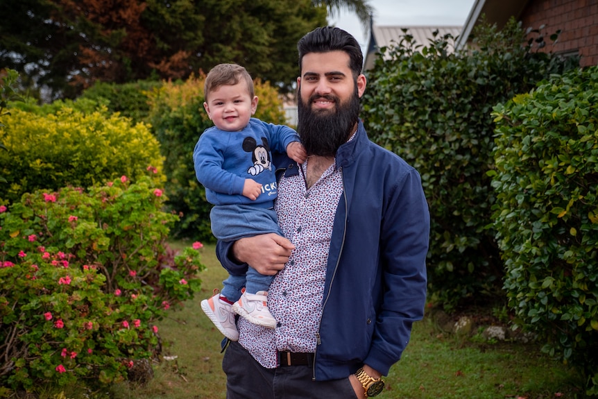 A smiling, dark-haired, bearded man holding a little boy in their front garden.