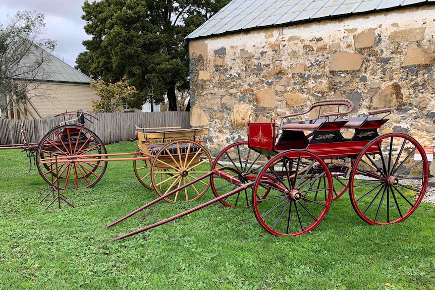 Three horse-drawn carriages lined up on grass outside an old sandstone building