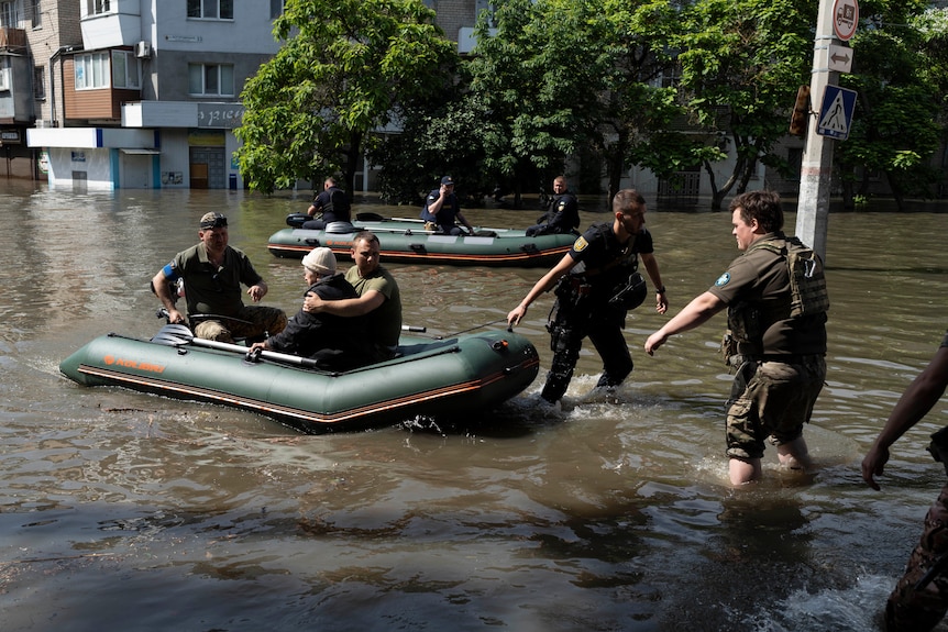 Rescuers use a small inflatable boat to resuce a woman, with two pulling her to safety through knee-high, brown floodwaters.