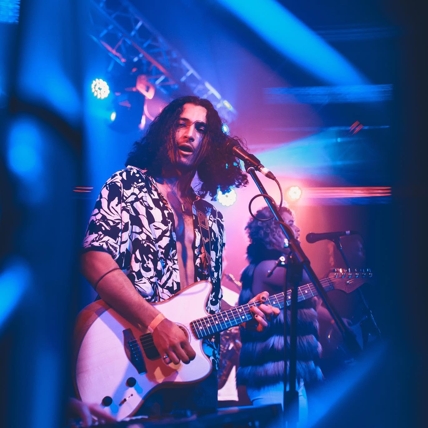 A man plays electronic guitar on a stage with neon lights around. 
