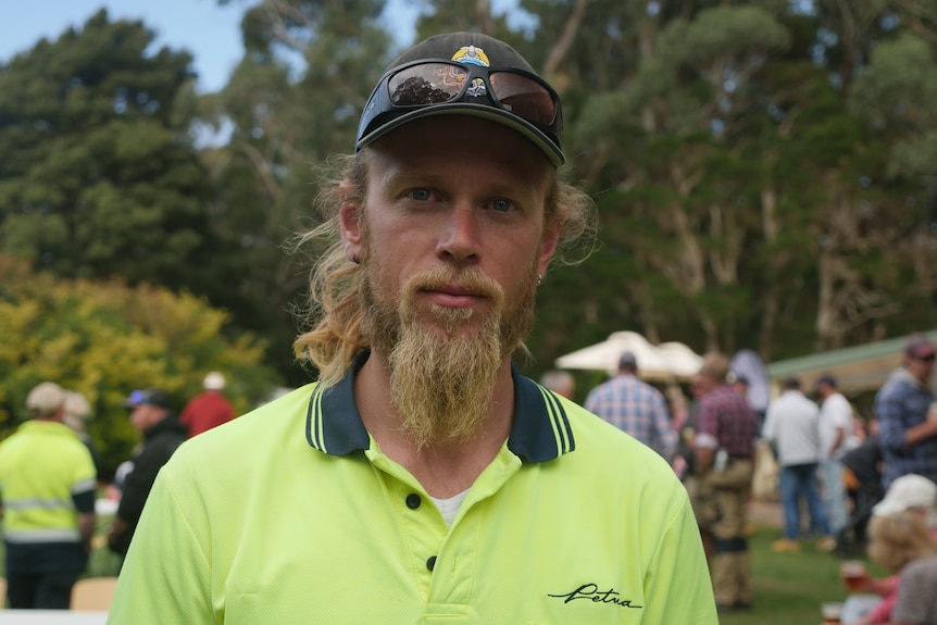 A man with long blond hair and beard, wearing a fluorescent yellow t-shirt and black cap and sunglasses on his head.
