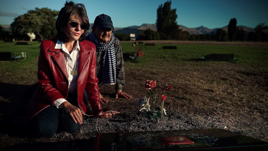 A couple sit at a gravestone with fake roses in the dirt