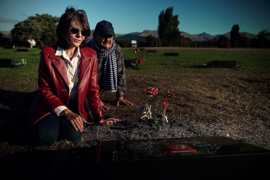 A couple sit at a gravestone with fake roses in the dirt