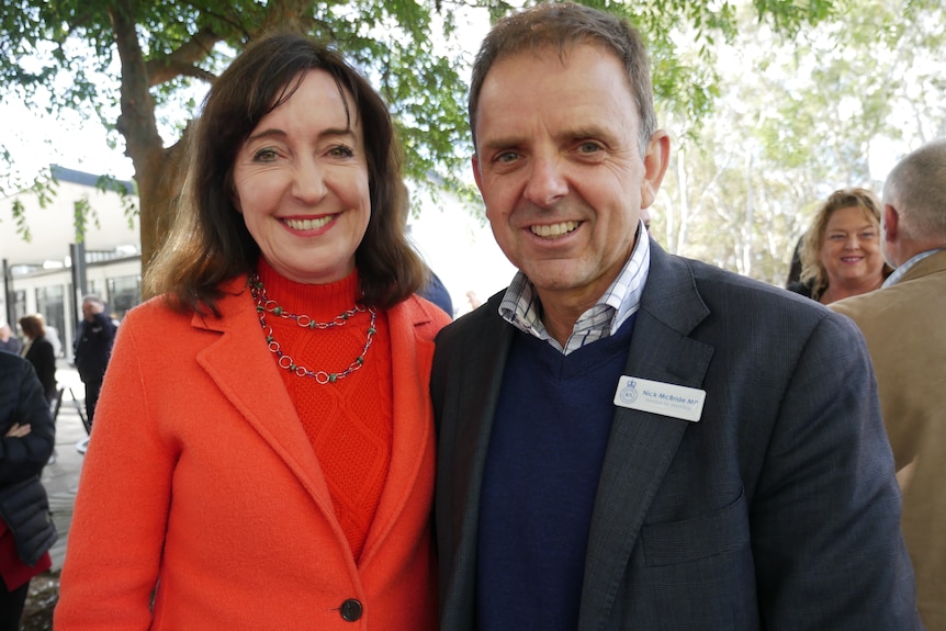 A brunette woman with an orange coat is pictured with a man wearing a checked blazer. They are smiling.