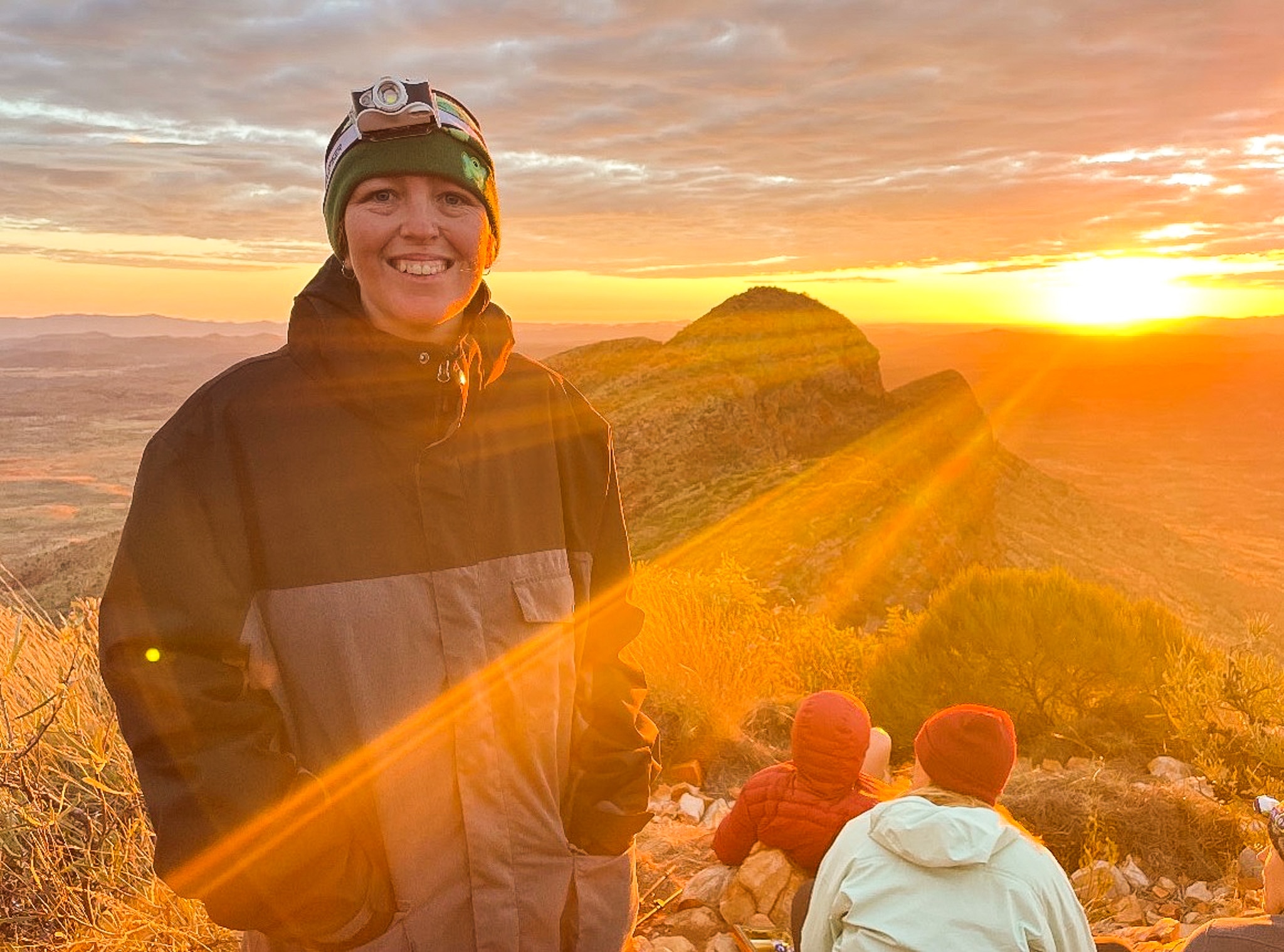 A woman wearing a beanie and puffer jacket smiles as the sun rises over a mountain.