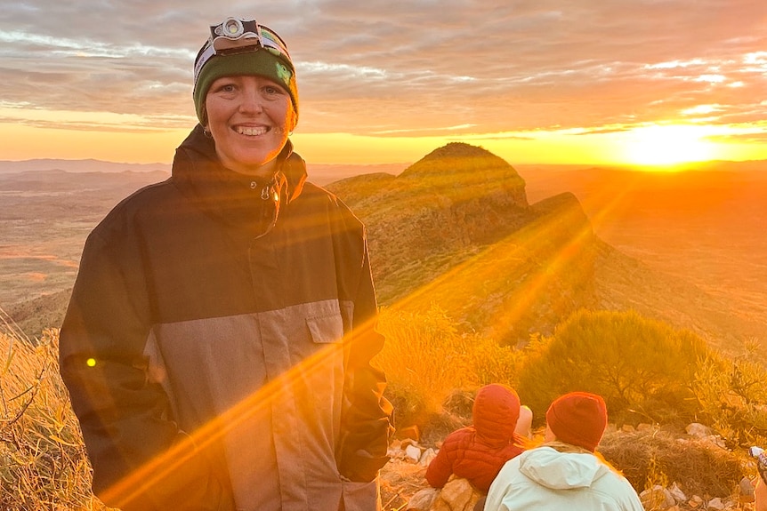 A woman wearing a beanie and puffer jacket smiles as the sun rises over a mountain.