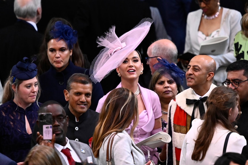 Katy Perry in a pink outfit and hat stands among guests at the coronation of King Charles III