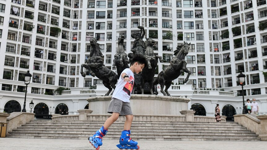 A boy skates in front of a residential complex, with a statue of a woman and horses out the front.