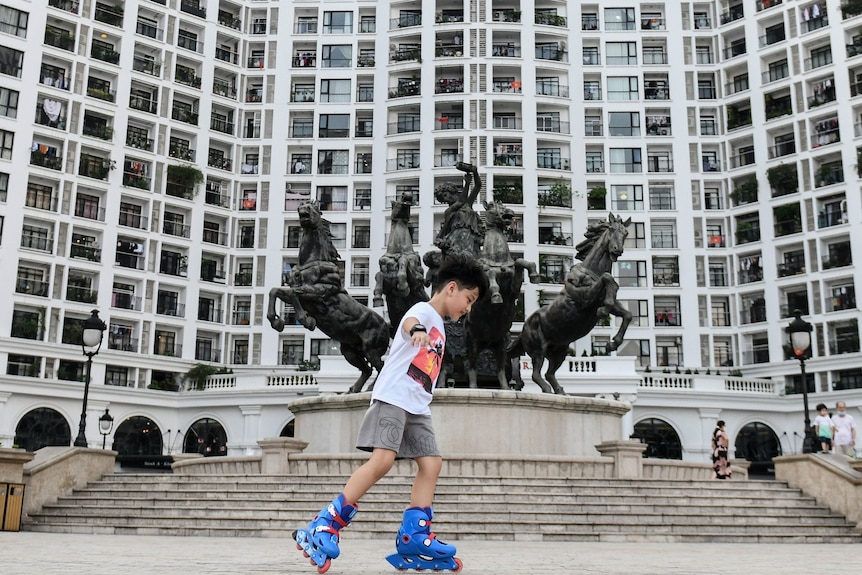 A boy skates in front of a residential complex, with a statue of a woman and horses out the front.