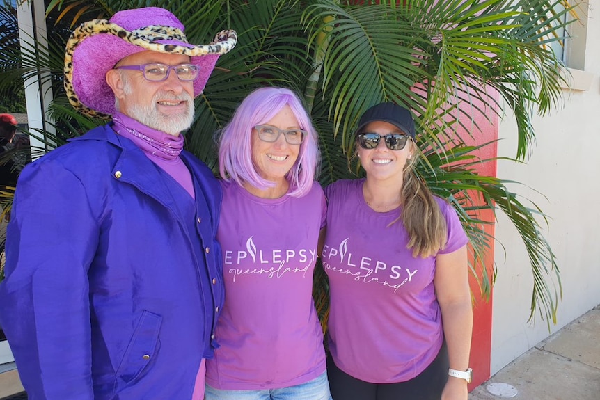 A man and two women stand dressed in purple with one of the woman with purple hair and the man in a dark purple jacket. 