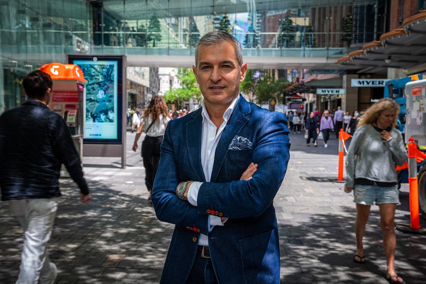 A man with grey hair, wearing a blue suit stands in a the CBD near shops, with his arms folded as he stares into the camera.