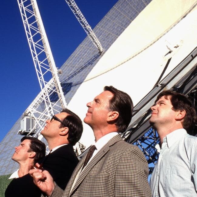 Sam Neill and two other cast members smile up at the Moon, sun on their faces.
