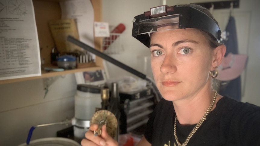 A woman with a neutral expression and blue eyes wearing a safety hard cap, she is holding a small tool with a sapphire on it