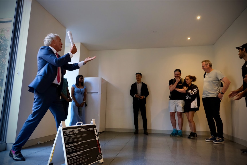 An auctioneer in a suit inside an apartment living room with six people watching. A young couple in a corner looks excited.