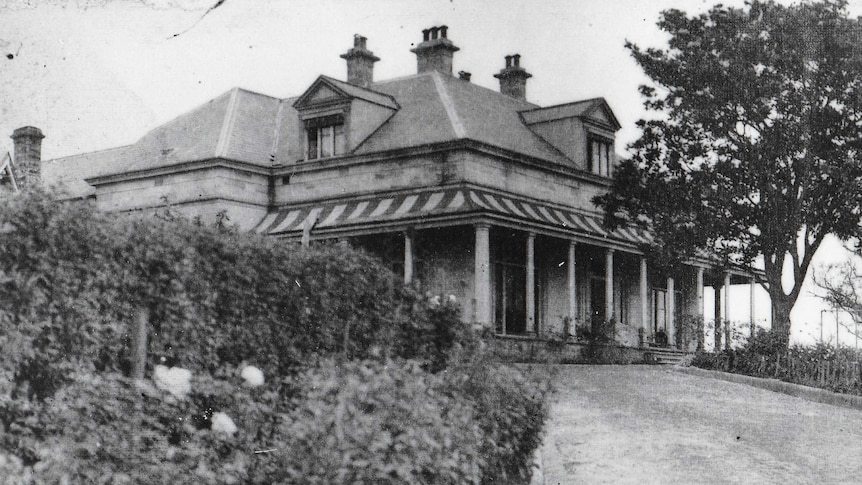 A black-and-white phot of a large house, lined with trees.