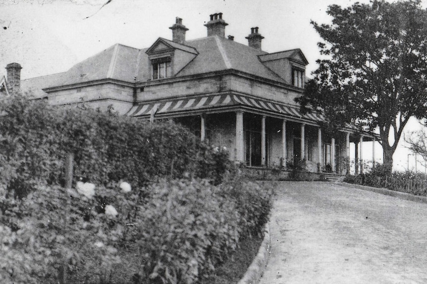 A black-and-white phot of a large house, lined with trees.